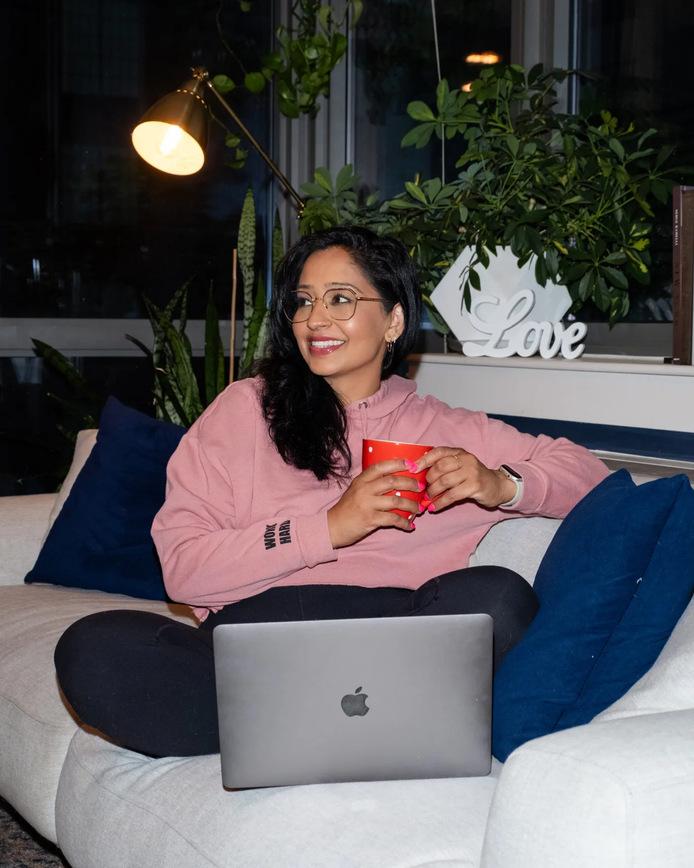 a women sitting cross legged on the couch with a mug in her hand in front of a laptop