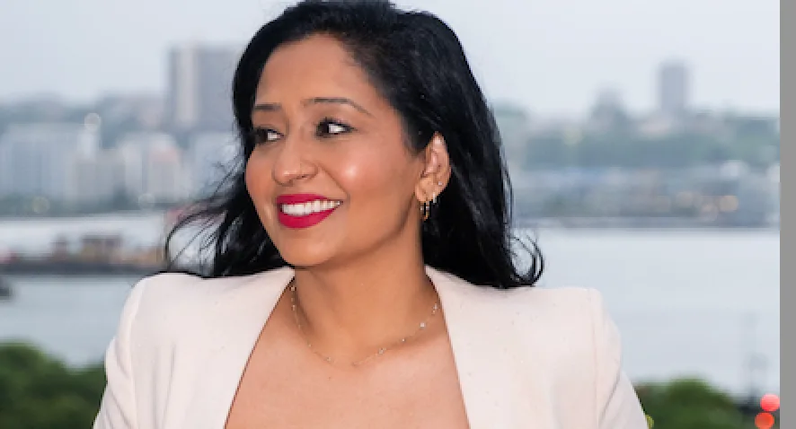 Professional woman with long dark hair and red lipstick wearing a beige blazer over a black top, sitting outdoors with a waterfront city skyline in the background