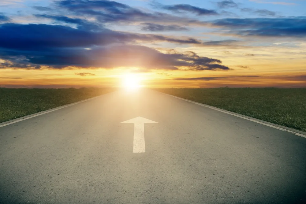 Asphalt road with white arrow sign goes to horizon. Sunshine on horizon and the road strigt away forward. Moving forward concept. Clear and positive perspectives ahead concept