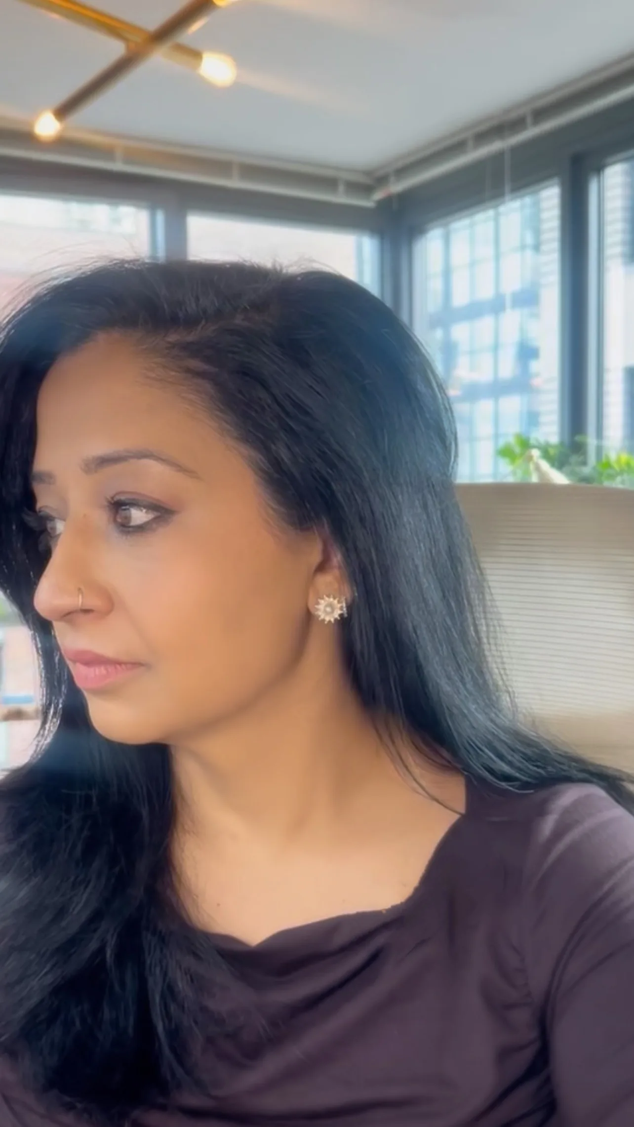 Professional woman with long dark hair and gold sunburst earrings looking thoughtfully to the side in a modern office setting with Professional woman with long dark hair and gold sunburst earrings looking thoughtfully to the side in a modern office setting with natural lighting and city viewsnatural lighting and city views