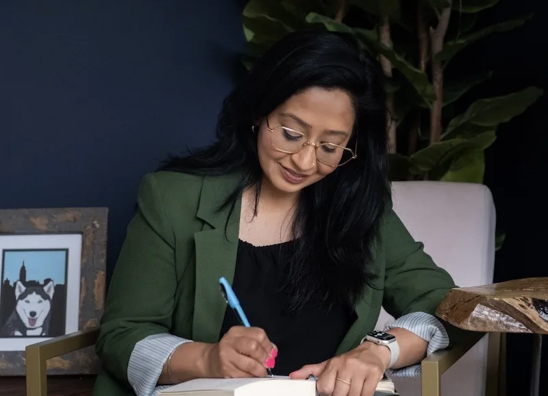 Professional woman in green blazer and glasses writing in notebook at wooden desk with plant and framed photo in background