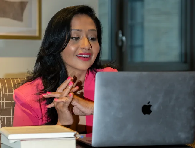 Woman in pink blazer gesturing while speaking and working on MacBook laptop in modern office setting
