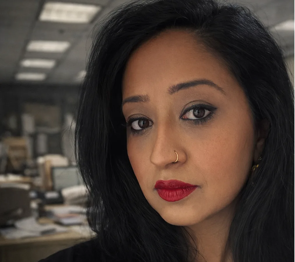 Close-up portrait of a professional woman with long dark hair and red lipstick, looking directly at the camera with a serious expression in a blurred office setting, with text overlay reading “Your worst job might be the best thing that ever happened.”