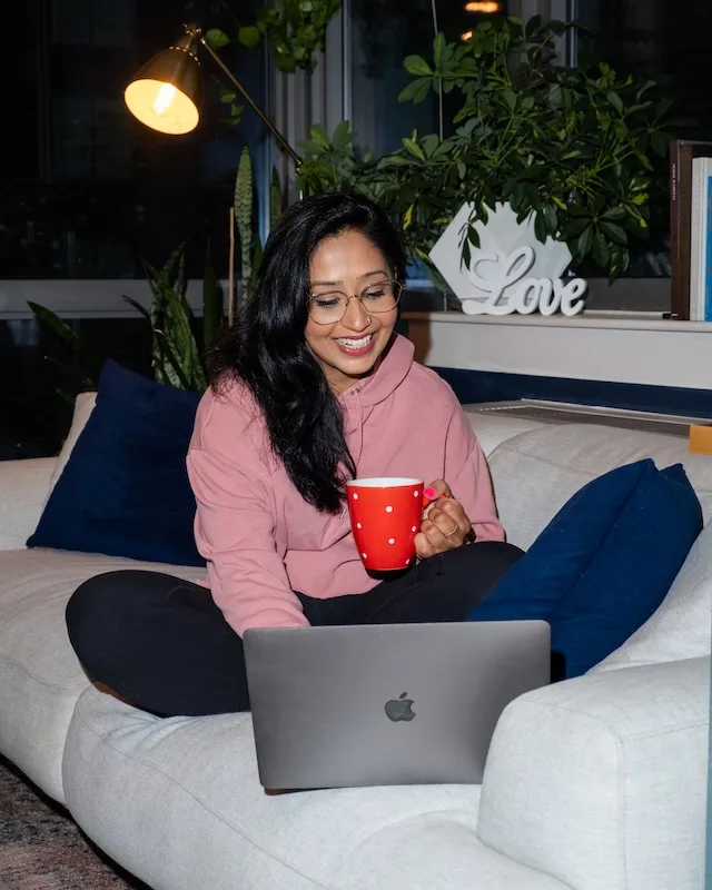 Professional woman with glasses and long dark hair wearing a pink sweatshirt, sitting cross-legged on a gray couch with blue pillows, holding a red mug while smiling. A MacBook laptop is open in front of her, with plants and warm lighting in the background creating a cozy home office environment.