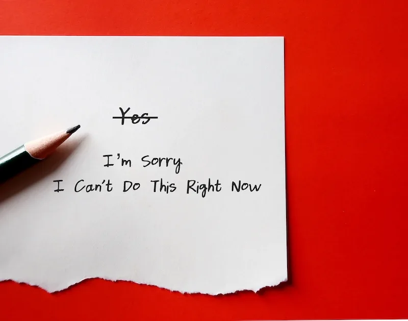 A handwritten note on white paper with the word "Yes" crossed out and the message "I'm sorry, I can't do this right now" written underneath, placed on a red background with a pencil pointing toward the text.